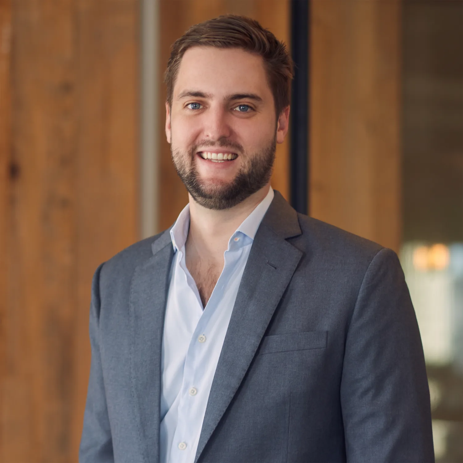 A person wearing a grey suit jacket over a light blue button-up shirt stands indoors near a wooden wall and glass partition.