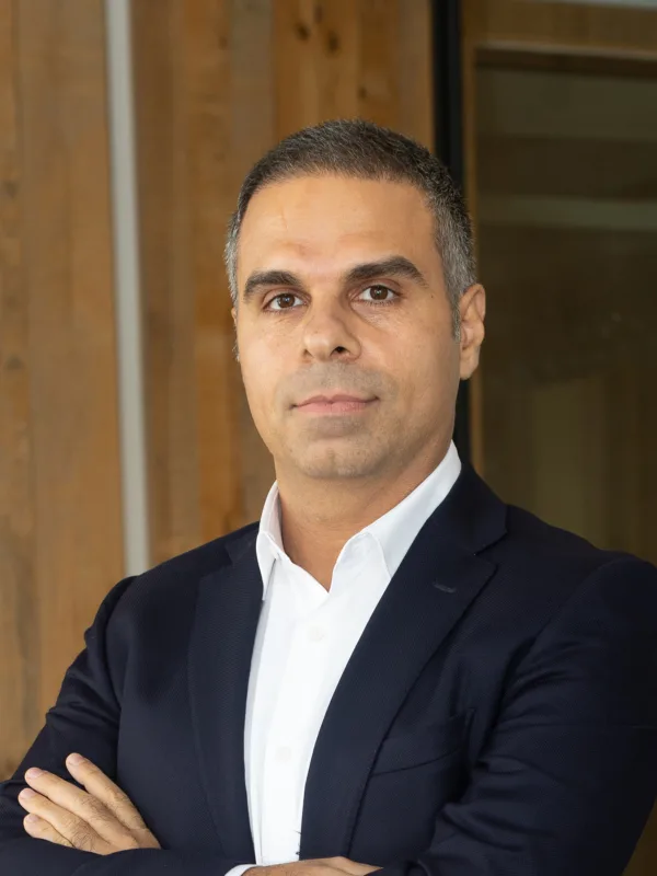 Fazel in a navy blue suit and white dress shirt stands with arms crossed in an office setting. The background includes wooden paneling and glass walls.