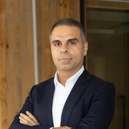 Fazel in a navy blue suit and white dress shirt stands with arms crossed in an office setting. The background includes wooden paneling and glass walls.