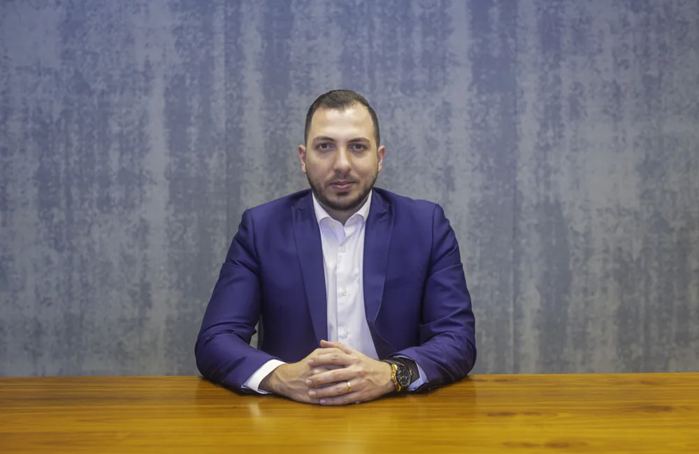 Mario Saab in a white shirt and blue jacket sat at a wooden table in front of a grey wall