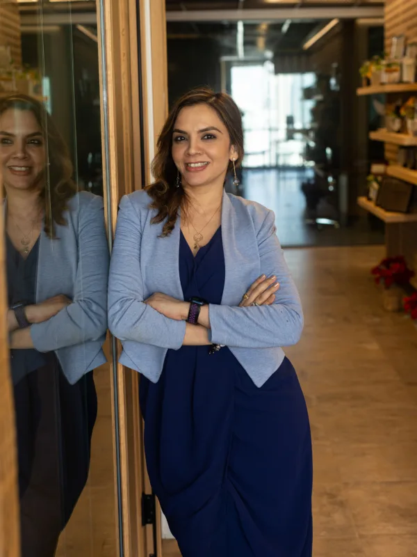 A person in a blue dress and light gray blazer stands indoors with arms crossed, leaning against a glass wall that reflects their figure. The hallway behind them is lined with shelves, plants, and framed items.