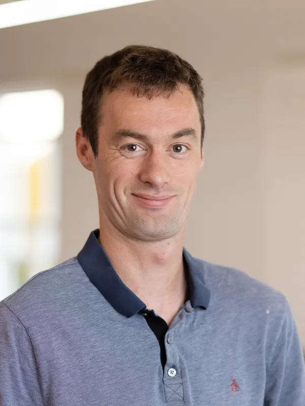 Head and shoulders image of Ben Marsh wearing a light blue polo shirt with a dark blue collar, with a blurred background of an office behind him