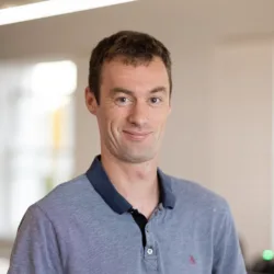 Head and shoulders image of Ben Marsh wearing a light blue polo shirt with a dark blue collar, with a blurred background of an office behind him