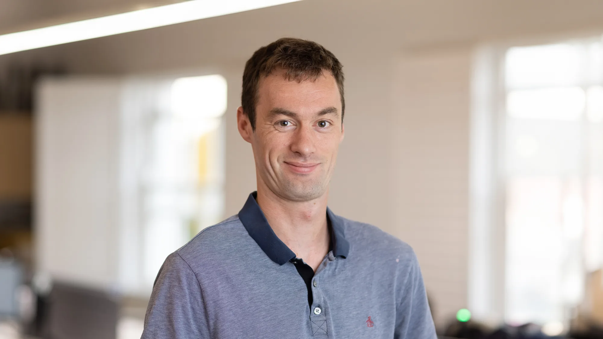 Head and shoulders image of Ben Marsh wearing a light blue polo shirt with a dark blue collar, with a blurred background of an office behind him