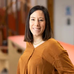 Ester smiling to camera in a long sleeve mustard top with the office behind