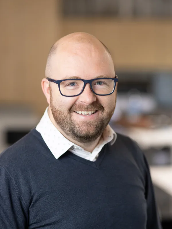 Gavin smiling to camera in a v-neck sweater and white open collar shirt with a blurred office backdrop