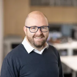 Gavin smiling to camera in a v-neck sweater and white open collar shirt with a blurred office backdrop