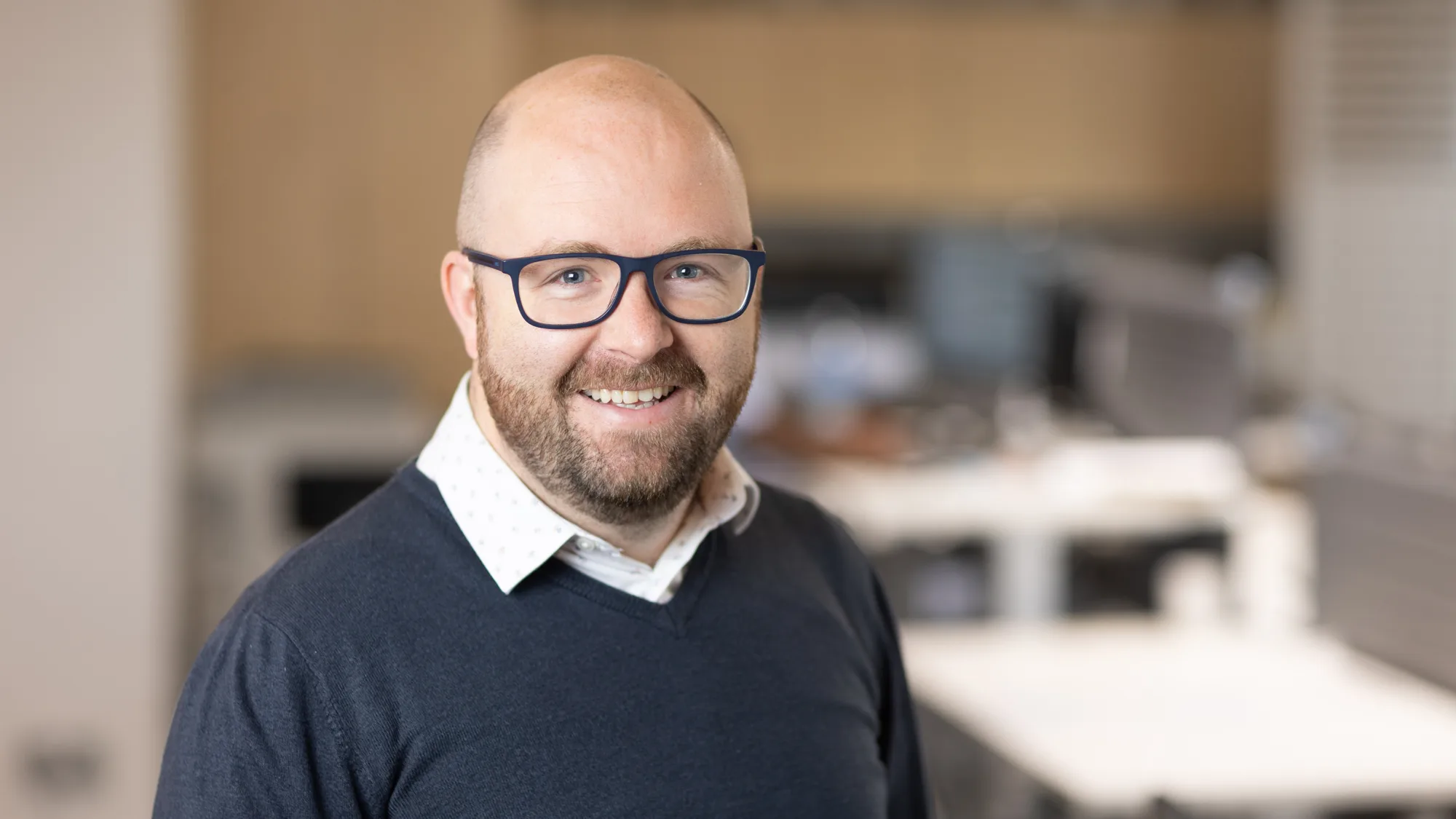 Gavin smiling to camera in a v-neck sweater and white open collar shirt with a blurred office backdrop