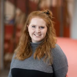 Hannah standing in a grey and black jumper with a soft focus view of the office behind