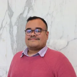Head and shoulders shot of Alex against a marble wall wearing glasses and a jumper