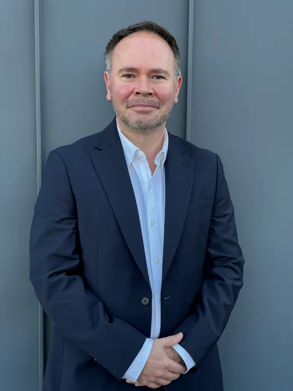 Portrait of an adult person standing against a plain grey wall covered with vertical panel lines, wearing a dark blazer over a light button-up shirt. Head and torso visible; hands are clasped in front.