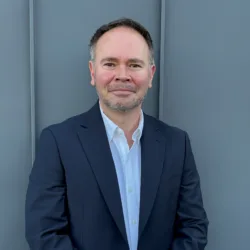 Portrait of an adult person standing against a plain grey wall covered with vertical panel lines, wearing a dark blazer over a light button-up shirt. Head and torso visible; hands are clasped in front.