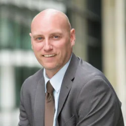 Graham sat in a pale shirt with grey suit jacket and brown tie smiling at the camera.
