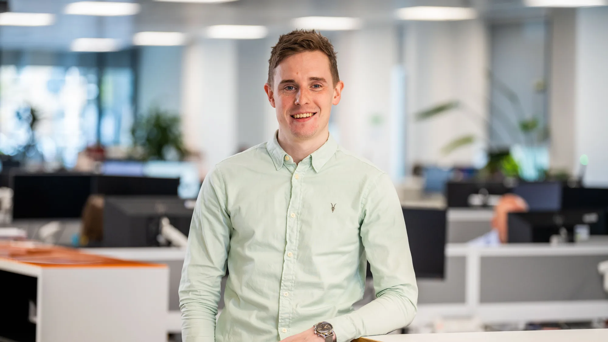 Jordan in a light green open collar shirt smiling to camera in a well lit office