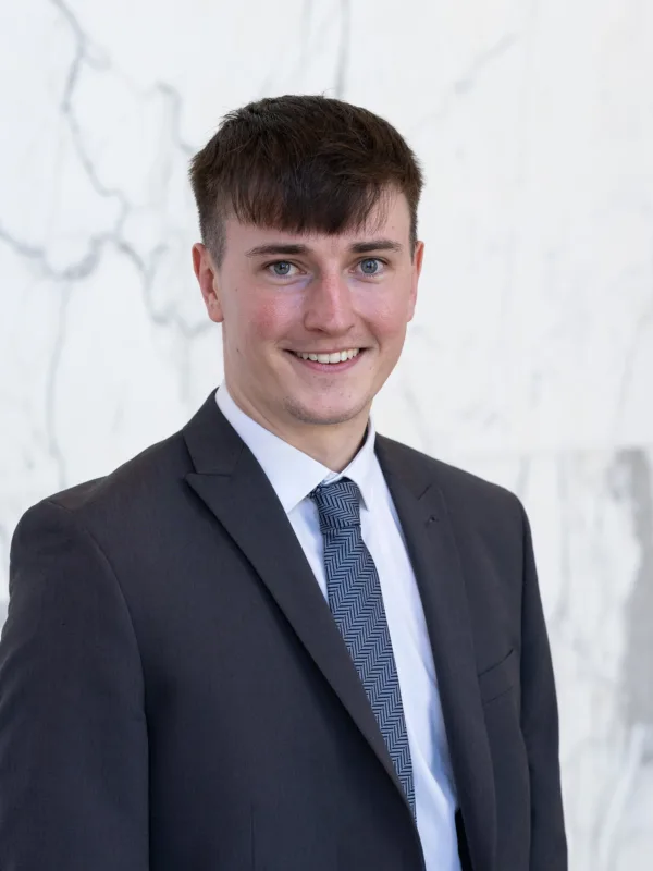 Jude stands in front of a light marble wall, wearing a dark textured suit jacket, white shirt, and patterned blue tie. Upper body portrait with neutral lighting and a clean background.