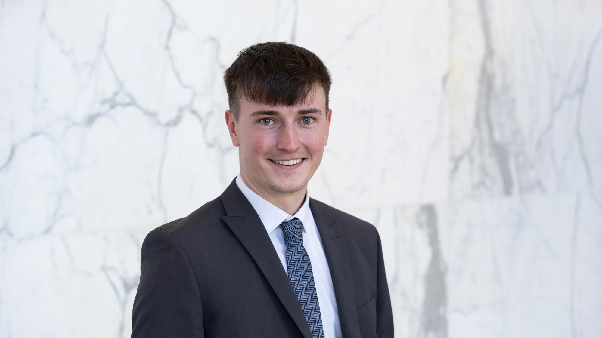 Jude stands in front of a light marble wall, wearing a dark textured suit jacket, white shirt, and patterned blue tie. Upper body portrait with neutral lighting and a clean background.