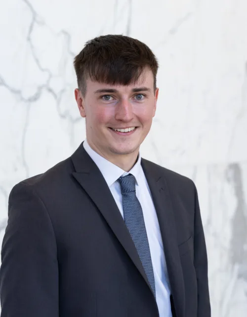 Jude stands in front of a light marble wall, wearing a dark textured suit jacket, white shirt, and patterned blue tie. Upper body portrait with neutral lighting and a clean background.