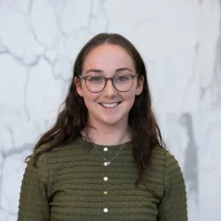 Person wearing an olive-green textured long-sleeve top with white and yellow buttons and a silver necklace with pendant, standing against a light grey marble wall with darker veining.