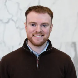 Head-and-shoulders portrait of a person wearing a dark quarter-zip sweater against a light marble-patterned background.