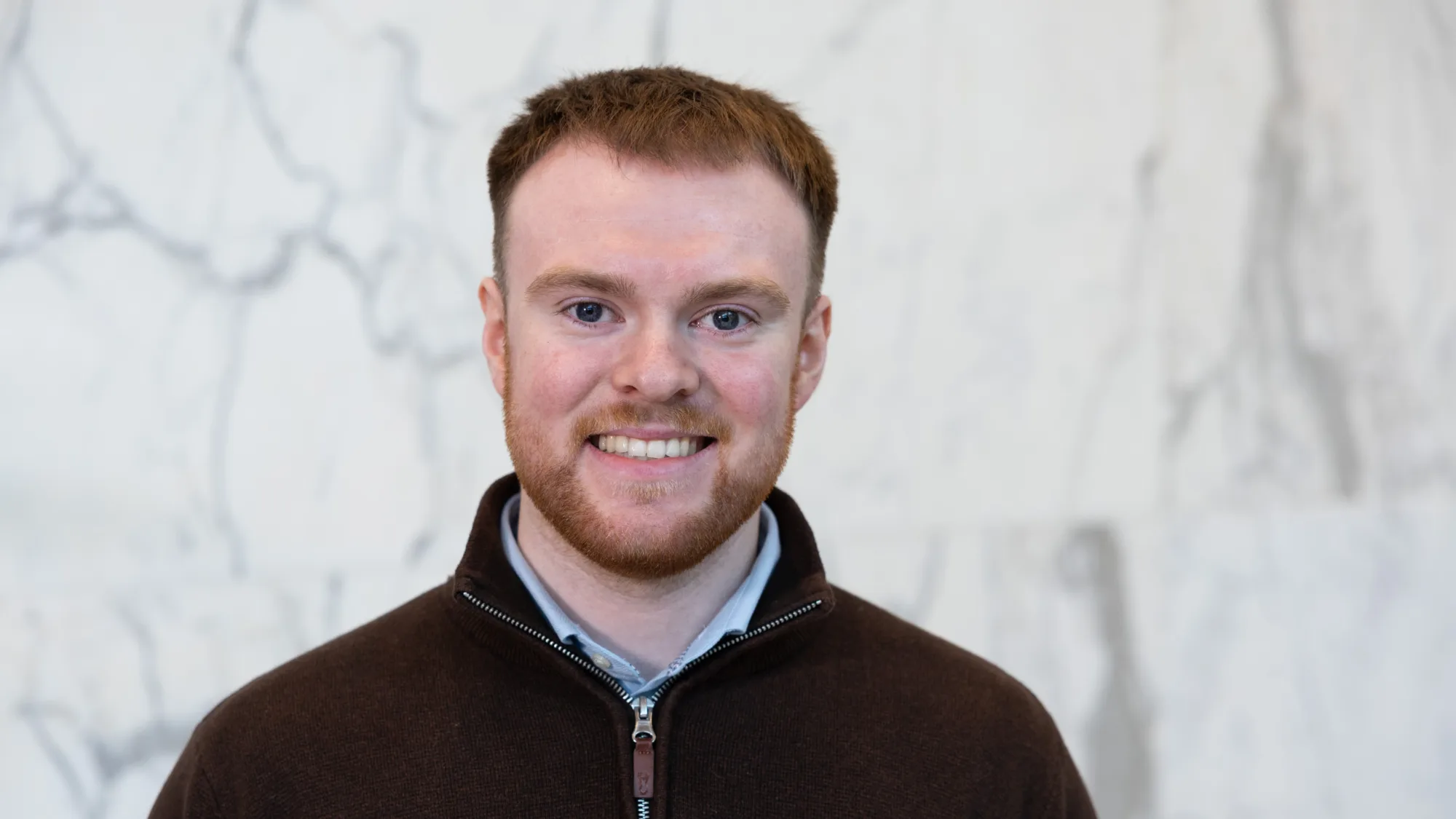 Head-and-shoulders portrait of a person wearing a dark quarter-zip sweater against a light marble-patterned background.