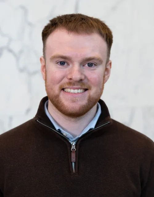 Head-and-shoulders portrait of a person wearing a dark quarter-zip sweater against a light marble-patterned background.
