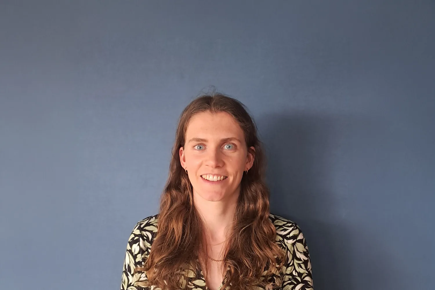 Head-and-shoulders portrait of a person with long wavy hair standing against a plain blue wall.