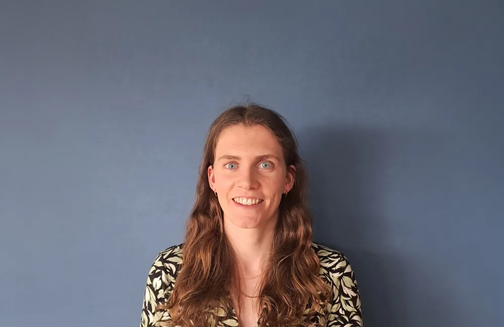 Head-and-shoulders portrait of a person with long wavy hair standing against a plain blue wall.
