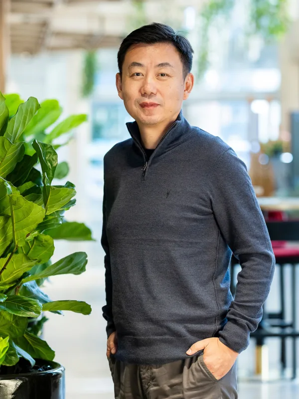 Dong stood in an open plan office with hands in pockets next to a tall green plant.