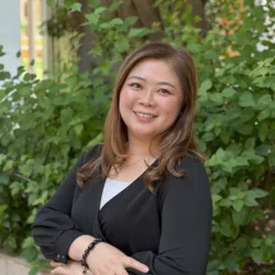 Person standing outdoors with arms crossed, wearing a black cardigan over a light top and bracelets on one wrist. Positioned in front of leafy shrubs and a tree, with a light-coloured building visible in the background.