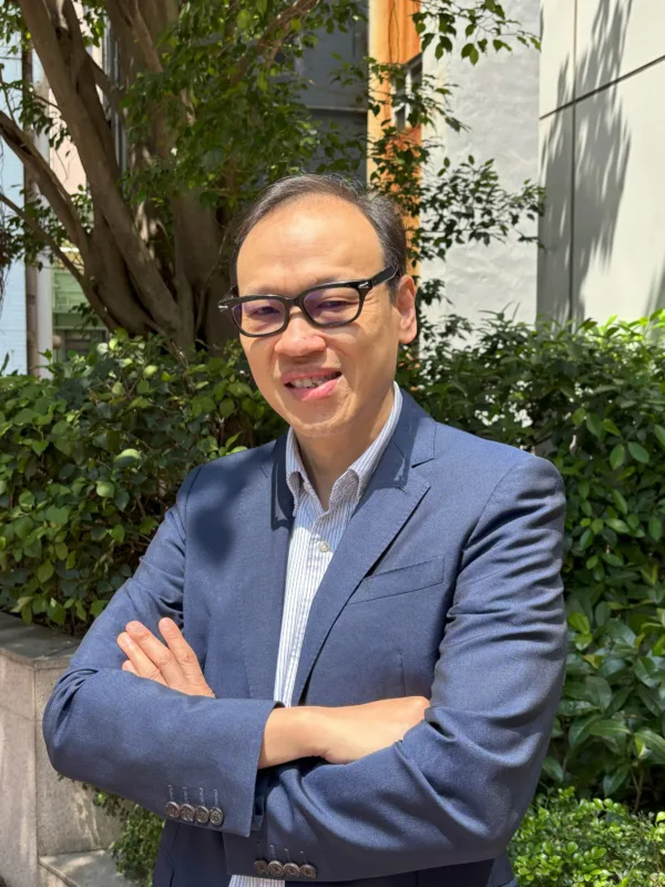 Person wearing glasses, a blue blazer, and a collared shirt stands outdoors with arms crossed. Green shrubs, a tree, and a light-coloured building wall form the background in bright sunlight.