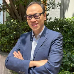 Person wearing glasses, a blue blazer, and a collared shirt stands outdoors with arms crossed. Green shrubs, a tree, and a light-coloured building wall form the background in bright sunlight.