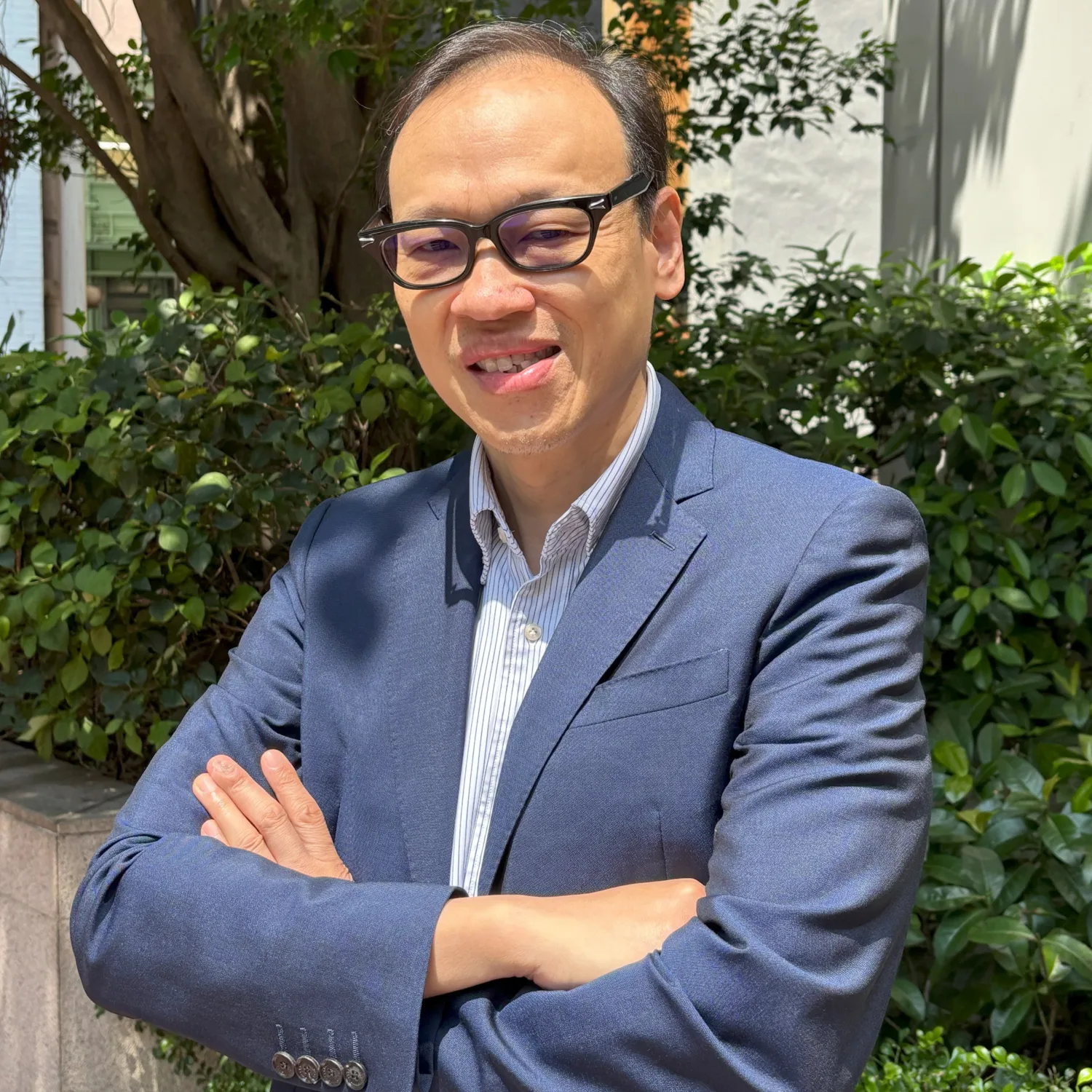Person wearing glasses, a blue blazer, and a collared shirt stands outdoors with arms crossed. Green shrubs, a tree, and a light-coloured building wall form the background in bright sunlight.