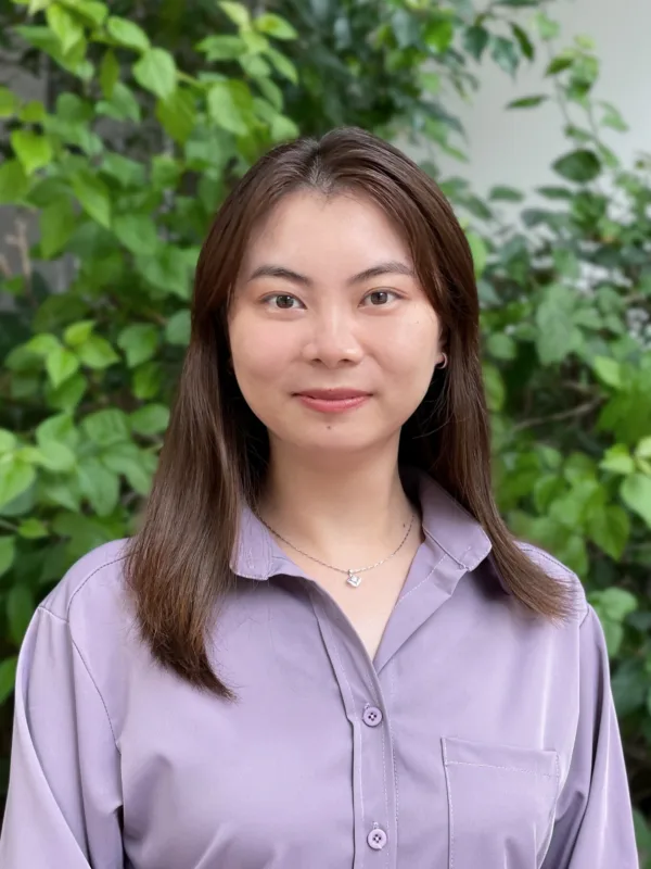 Ida in a lilac open collar shirt smiling to camera outside with a leafy back drop