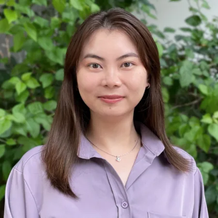 Ida in a lilac open collar shirt smiling to camera outside with a leafy back drop