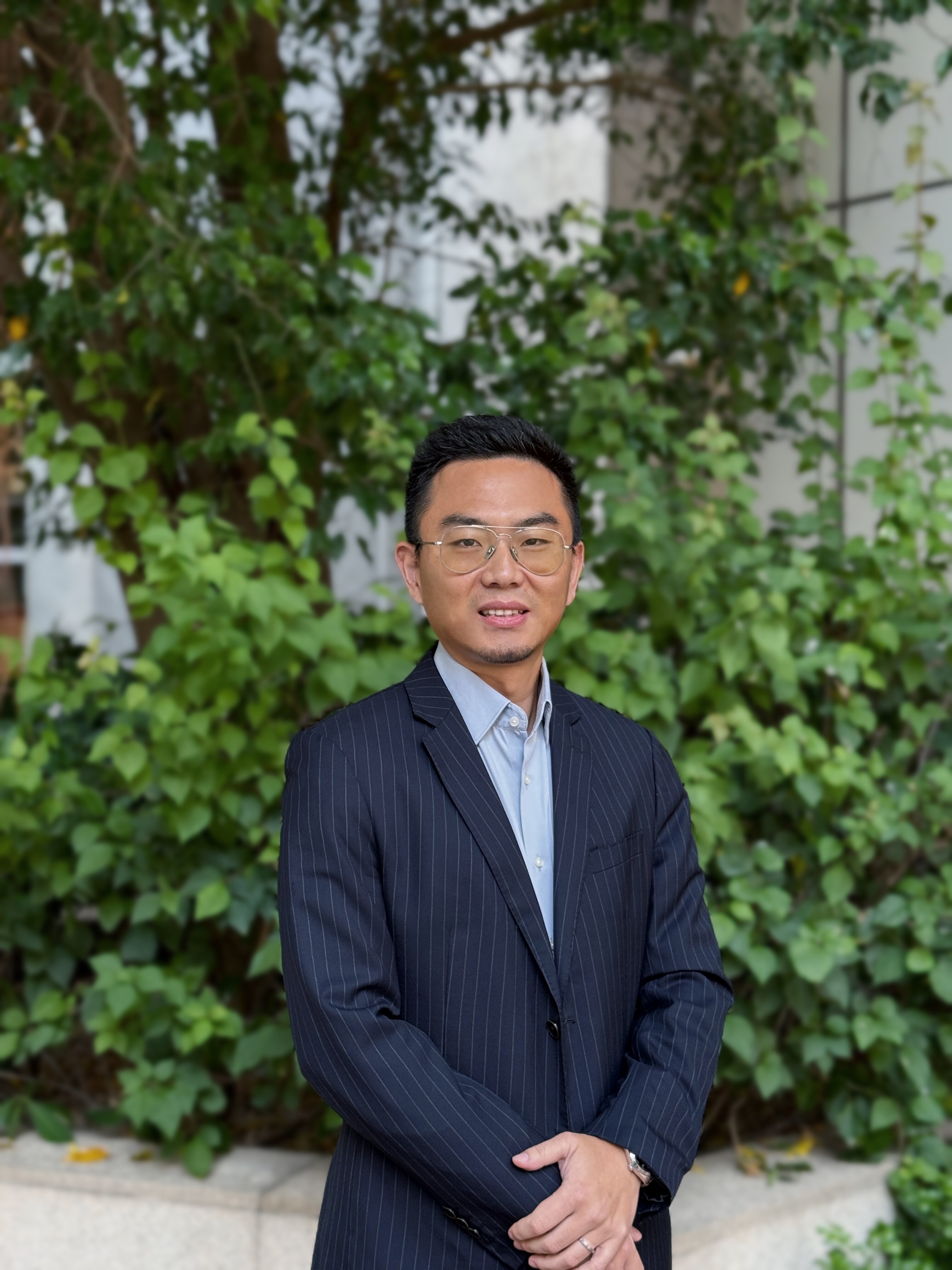 Kevin in a light blue shirt and dark suit in front of a leafy green border