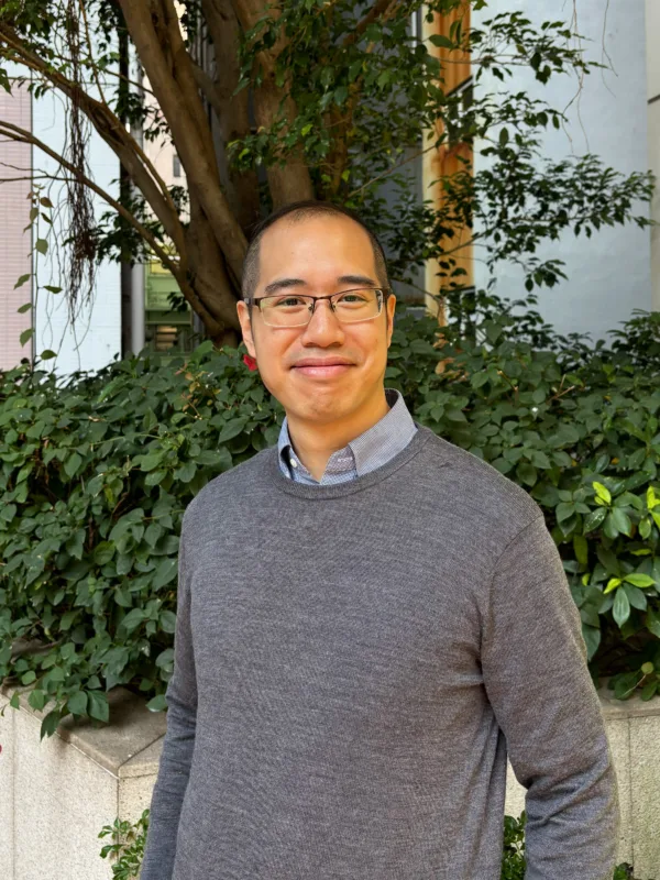 Person standing outdoors in front of dense green shrubs and a tree, wearing a grey textured sweater over a collared shirt. A low stone planter and a light-coloured building are visible in the background.