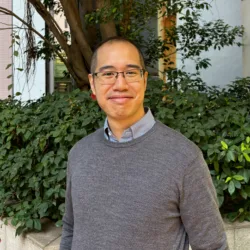Person standing outdoors in front of dense green shrubs and a tree, wearing a grey textured sweater over a collared shirt. A low stone planter and a light-coloured building are visible in the background.