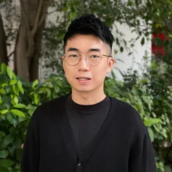Pete wearing a dark round neck tee and cardigan outside in front of greenery
