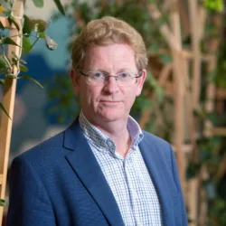 Alan Fogarty in London office reception in a suit witch wooden terrace panelling for plants in background