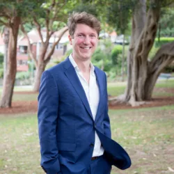 Alan wearing a white open collar shirt and blue suit in a suburban park