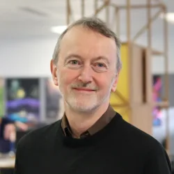 Alasdair smiling and looking at the camera in a brown shirt and black sweater in an office space