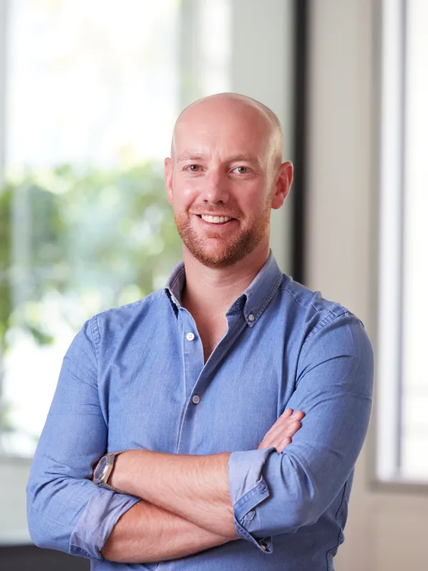 Andrew in an open collar blue shirt with arms folded, smiling to camera in front of the office window