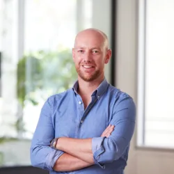 Andrew in an open collar blue shirt with arms folded, smiling to camera in front of the office window