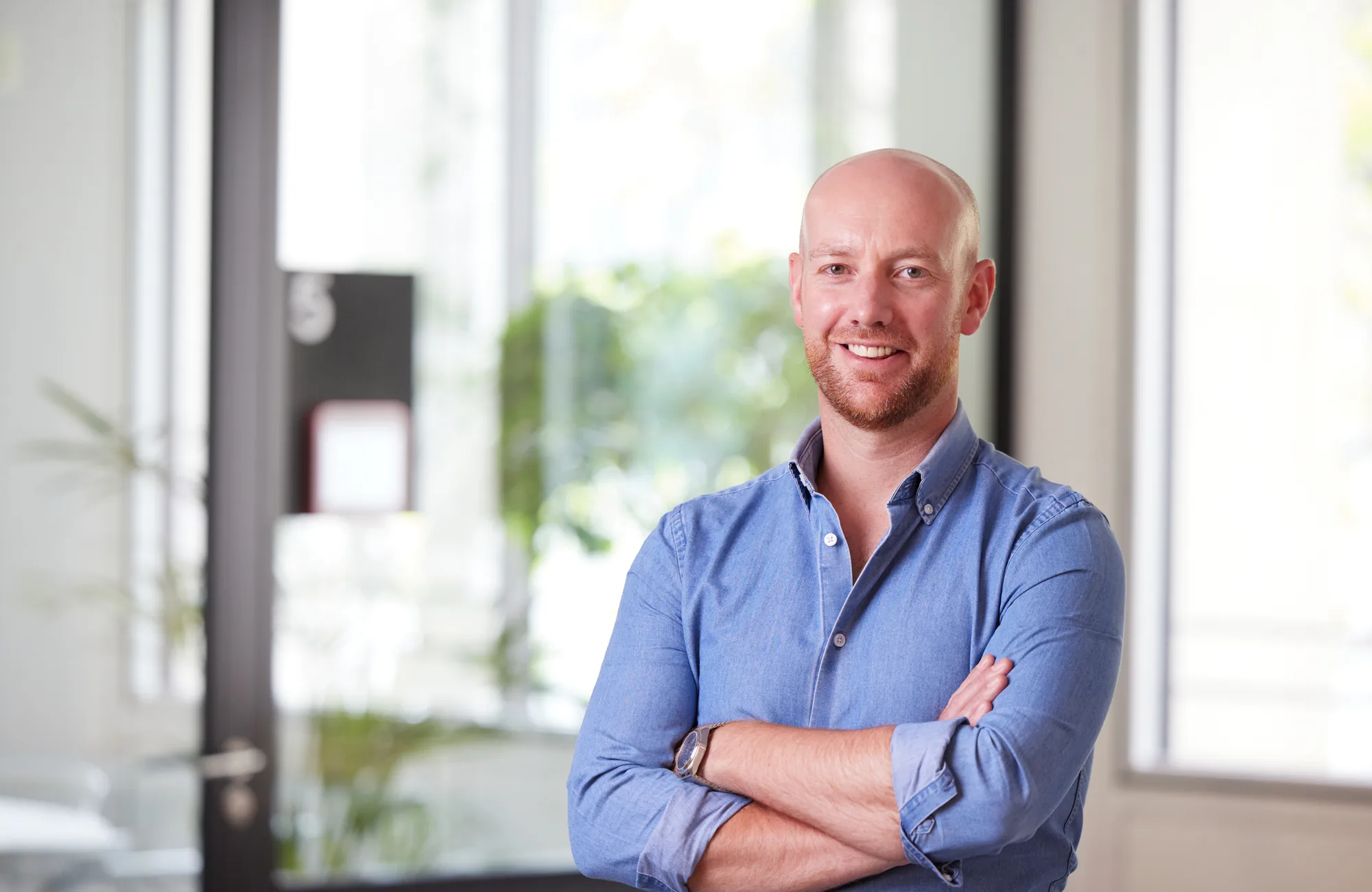 Andrew in an open collar blue shirt with arms folded, smiling to camera in front of the office window