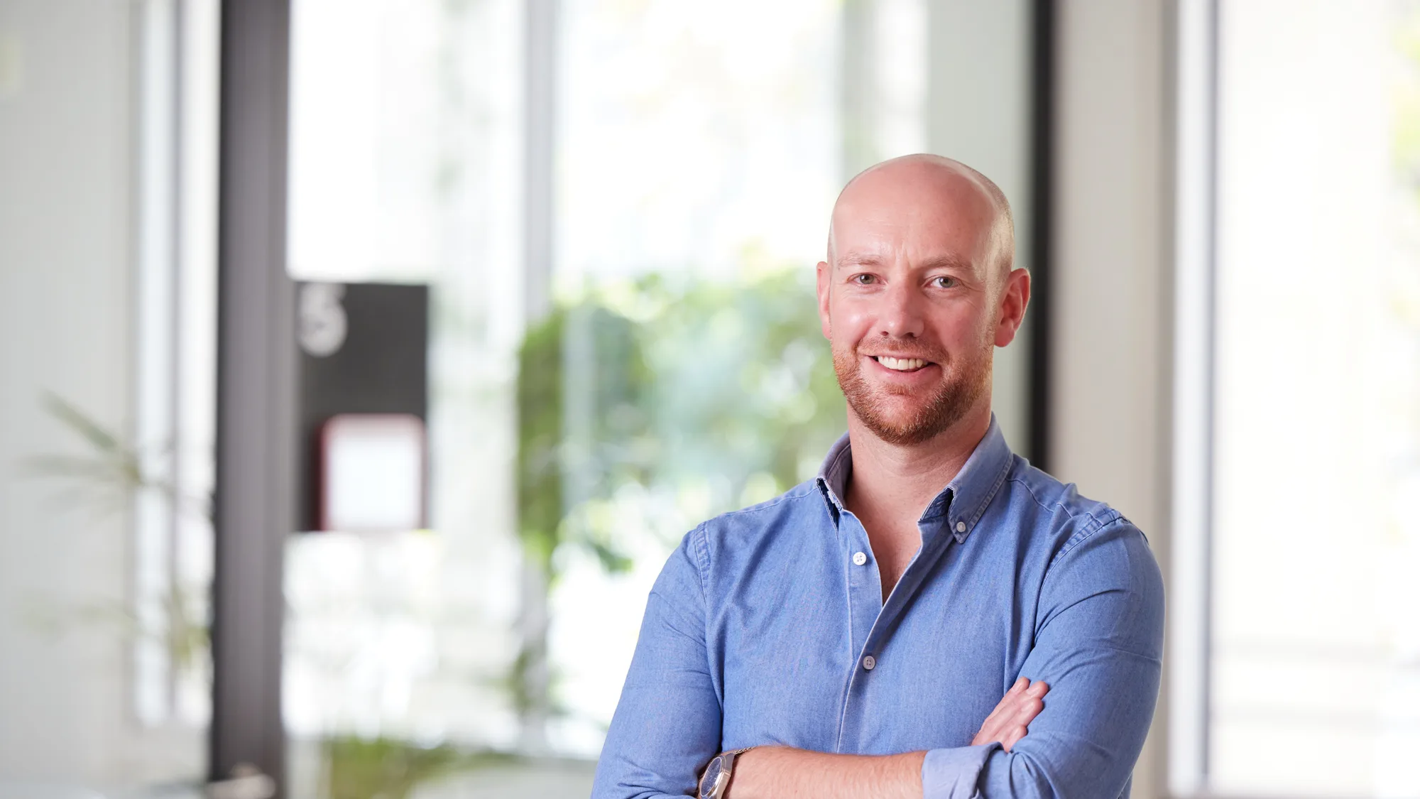 Andrew in an open collar blue shirt with arms folded, smiling to camera in front of the office window