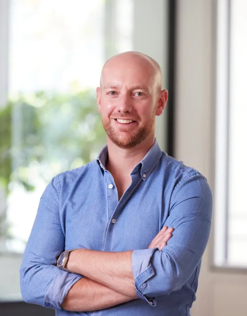 Andrew in an open collar blue shirt with arms folded, smiling to camera in front of the office window