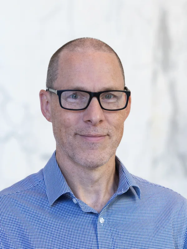 Head and shoulders image of Andrew stood in front of a white marble wall with a plant in the corner wearing a textured blue shirt