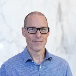 Head and shoulders image of Andrew stood in front of a white marble wall with a plant in the corner wearing a textured blue shirt