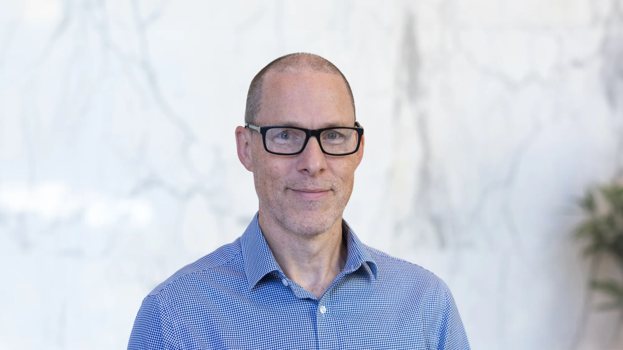Head and shoulders image of Andrew stood in front of a white marble wall with a plant in the corner wearing a textured blue shirt