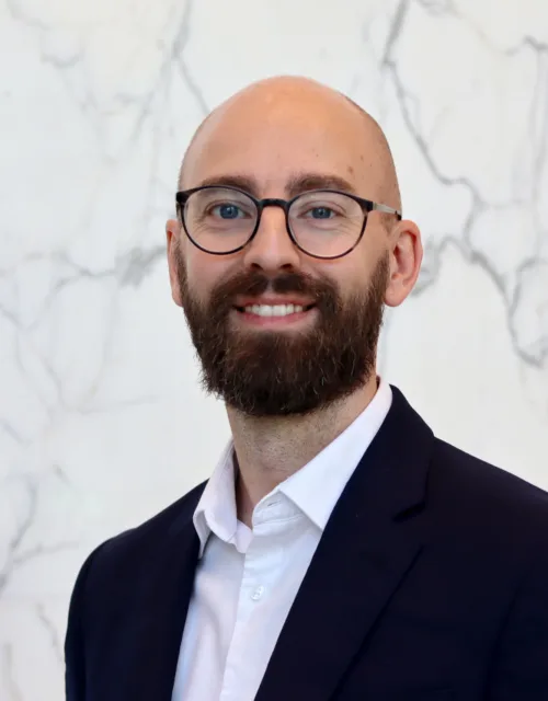 Andy in a dark suit jacket and white dress shirt stands in front of a white marble wall with grey veining.
